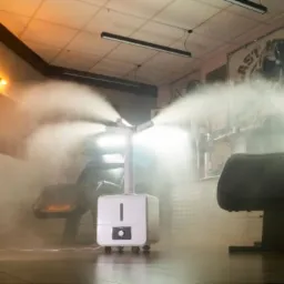 Biały, przenośny zamgławiacz rozpyla dezynfekującą mgłę w salonie fryzjerskim, z widocznymi fotelami fryzjerskimi i dekoracyjnym oświetleniem ściennym.