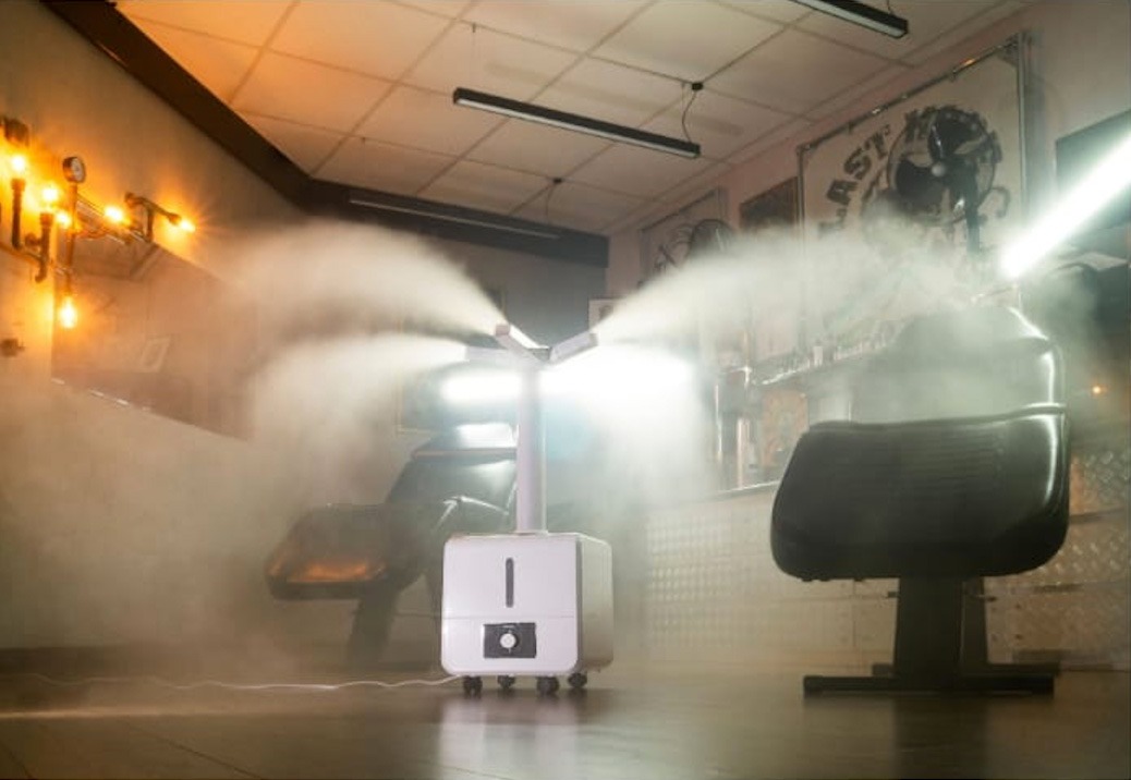 Biały, przenośny zamgławiacz rozpyla dezynfekującą mgłę w salonie fryzjerskim, z widocznymi fotelami fryzjerskimi i dekoracyjnym oświetleniem ściennym.