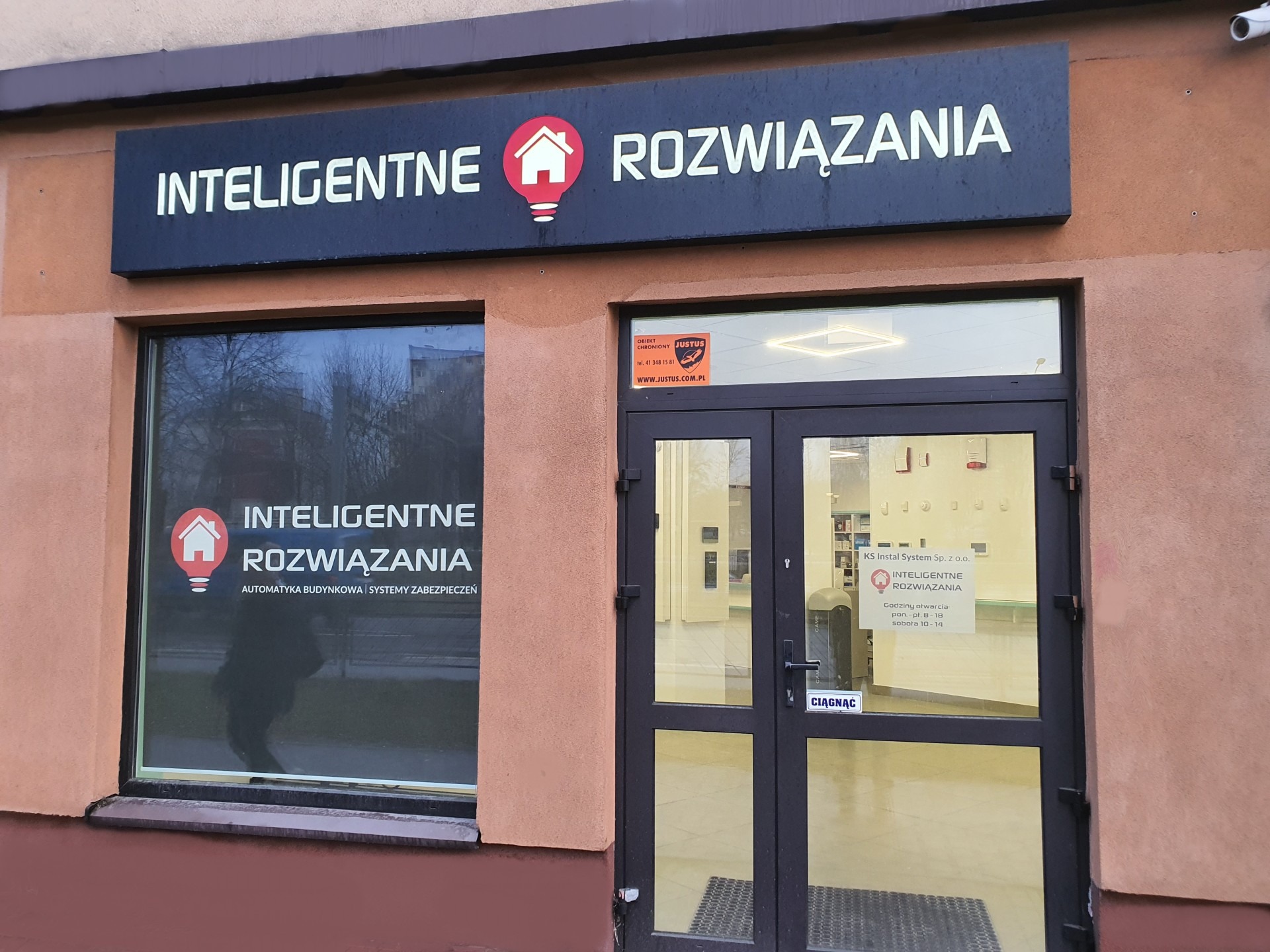Fasada budynku z szyldem 'Inteligentne Rozwiązania' z symbolem domu w żarówce, okno z naklejką o tej samej treści, oraz drzwi wejściowe z naklejką informującą o godzinach otwarcia i ochronie obiektu.