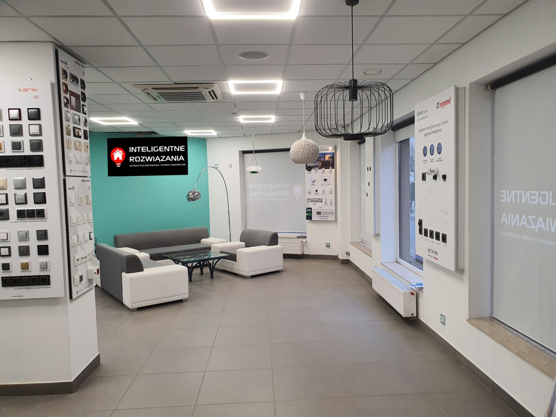 Salon ekspozycyjny inteligentnych rozwiązań domowych z ekspozytorami włączników Kariik, systemem Legrand Netatmo i strefą wypoczynkową z sofami.