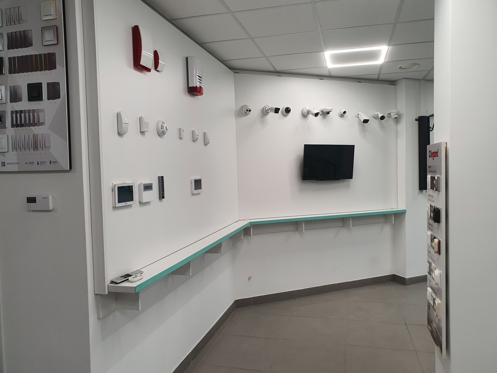 Ekspozycja różnorodnych elementów wyposażenia domu, w tym panel z włącznikami, czujniki, kamery monitoringu oraz panel sterowania, umieszczonych na białych ścianach w przestronnym wnętrzu sklepu...