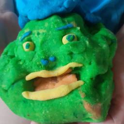 Kuferek Niespodzianek - Zielona postać z plasteliny z niebieskim kapeluszem, żółtymi oczami i ustami, trzymana w dłoniach dziecka.