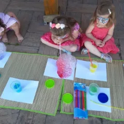 Trzy dziewczynki siedzą na macie i tworzą kolorowe bańki z płynu za pomocą słomek, widoczne kubki z płynem, kartki papieru i opakowanie pisaków.