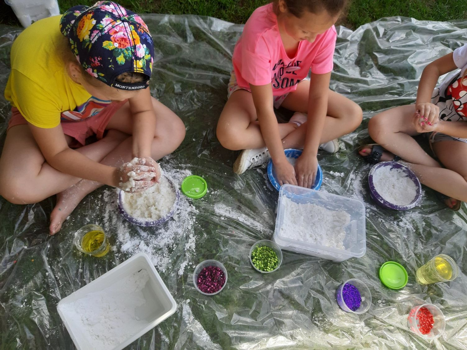 Trzy dziewczynki siedzą na folii ochronnej na trawie, mieszając składniki do sensorycznej zabawy, w tym biały proszek, brokat i olej, w miskach i pojemnikach.