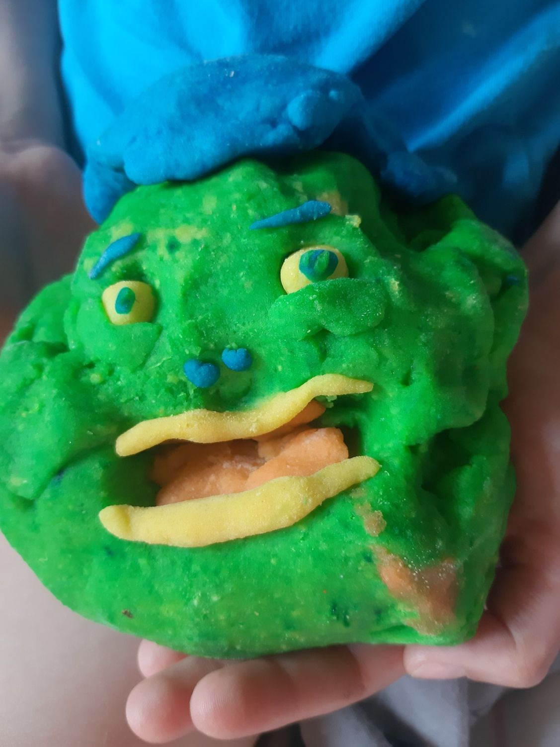 Zielona postać z plasteliny z niebieskim kapeluszem, żółtymi oczami i ustami, trzymana w dłoniach dziecka.