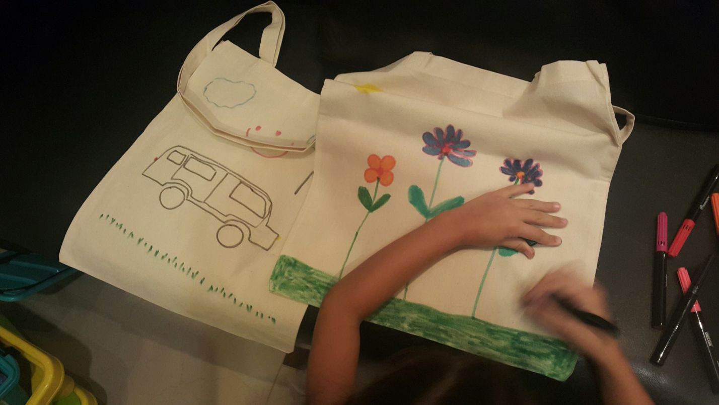 Dziecko koloruje flamastrem bawełnianą torbę z narysowanymi kwiatami; obok leży druga torba z rysunkiem kampera.