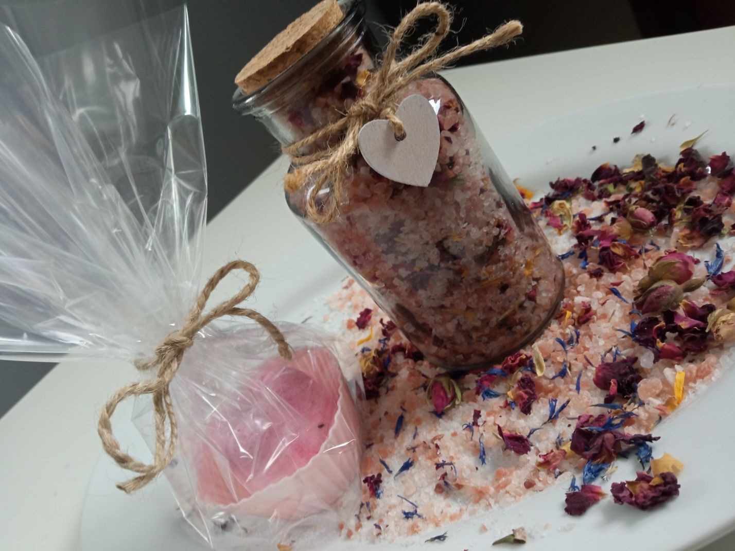 Ręcznie robione mydełko w celofanie i słoiczek z solą do kąpieli z dodatkiem suszonych kwiatów, ozdobione sznurkiem jutowym i serduszkiem, na białym talerzu.