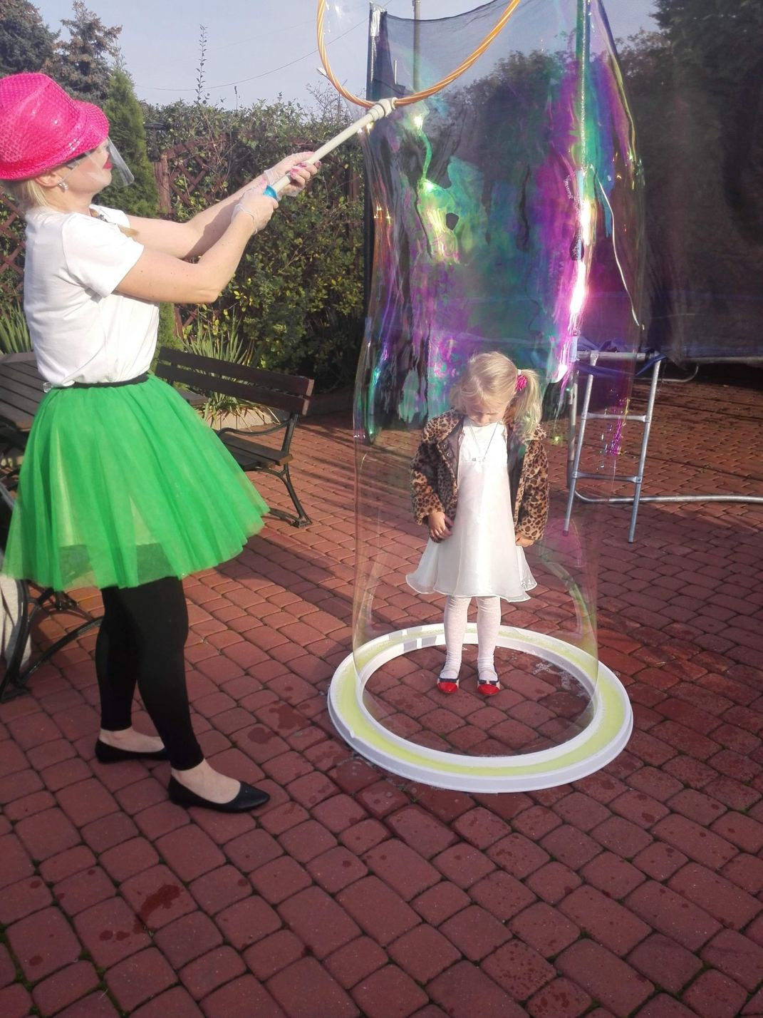 Dziewczynka w białej sukience i czerwonych butach stoi wewnątrz gigantycznej bańki mydlanej, tworzonej przez kobietę w różowym kapeluszu, zielonej spódnicy tiulowej i białej koszulce na tle...
