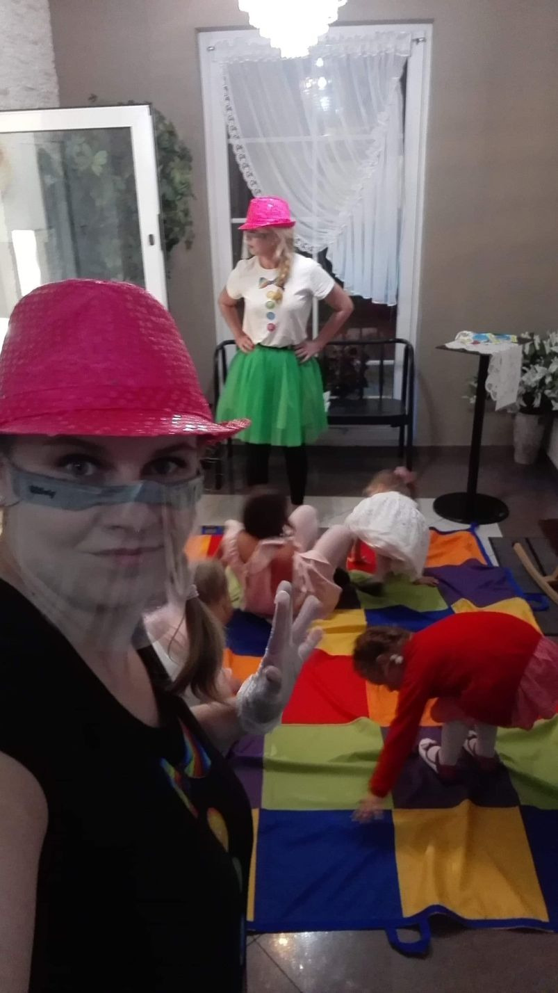 Animator w czarnej koszulce z tęczowym motywem i osłoną na twarzy, prowadzi animacje dla dzieci na kolorowej macie w kratkę, w tle drugi animator w różowym kapeluszu i zielonej spódniczce.