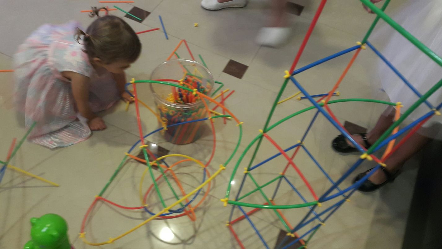 Dziewczynka w sukience bawi się kolorowymi rurkami konstrukcyjnymi, tworząc geometryczne budowle. Obok stoi przezroczysty pojemnik z dodatkowymi elementami, a na podłodze leży zielona zabawka...