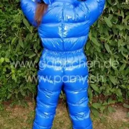 Osoba w błyszczącym, niebieskim kombinezonie puchowym, stojąca tyłem na trawie przed zielonym krzewem. Widoczny fragment brązowych włosów spod kaptura.