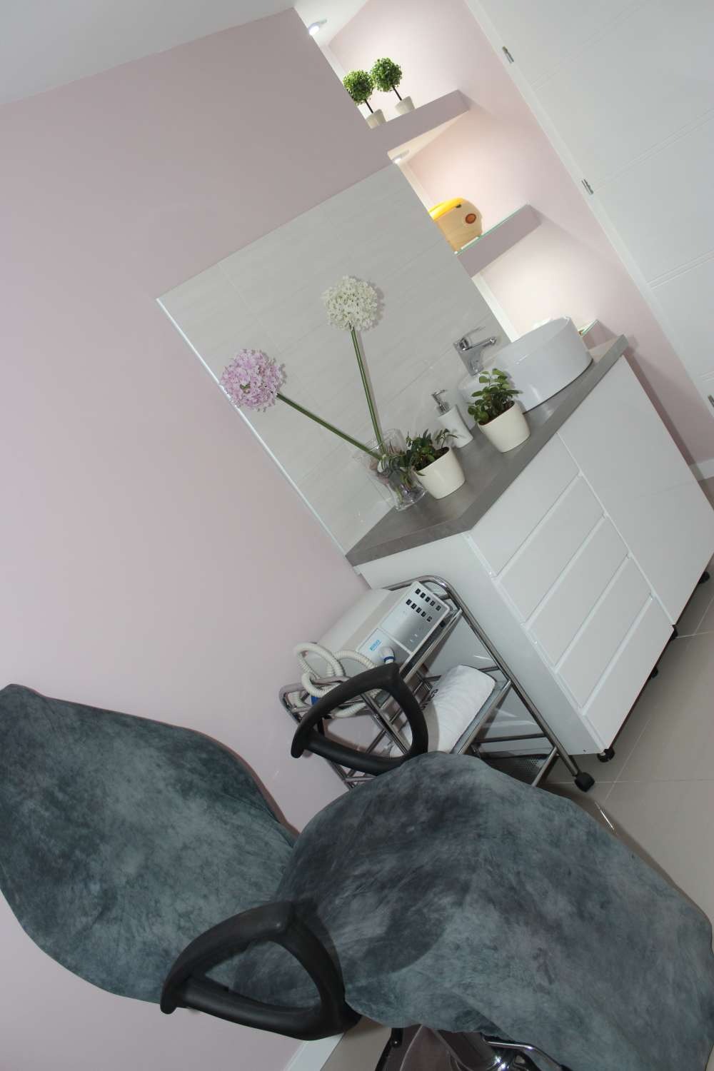 Wnętrze gabinetu: fotel zabiegowy, biała szafka z umywalką, kwiaty w wazonie i sprzęt kosmetyczny na wózku.