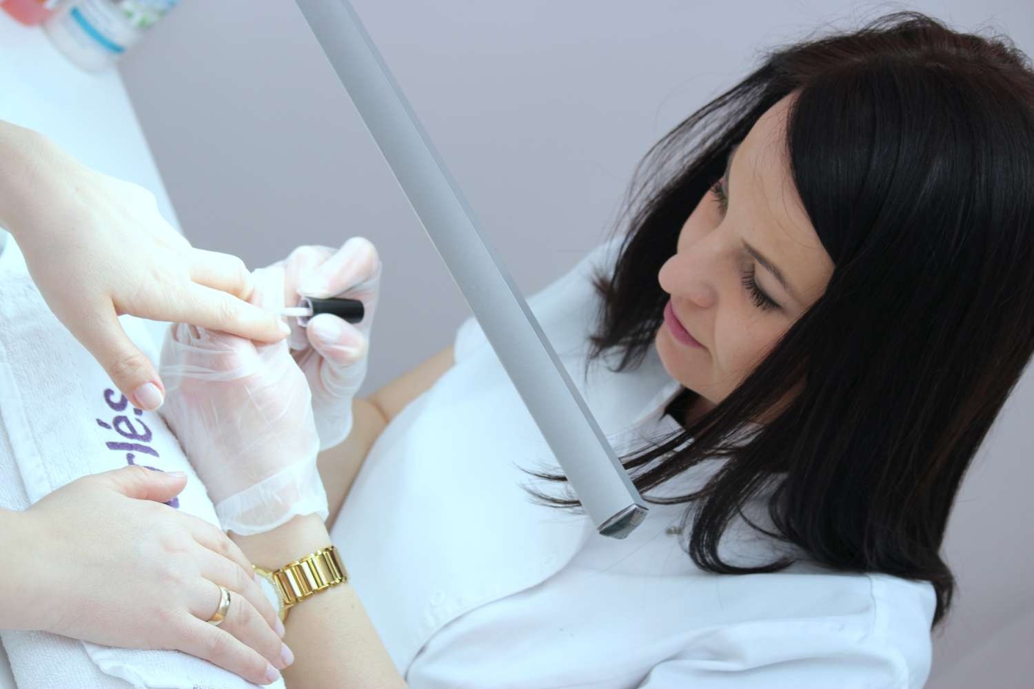 Kobieta w białym uniformie, w rękawiczkach, precyzyjnie maluje paznokcie klientce pod lampą, widoczny złoty zegarek na ręce klientki.