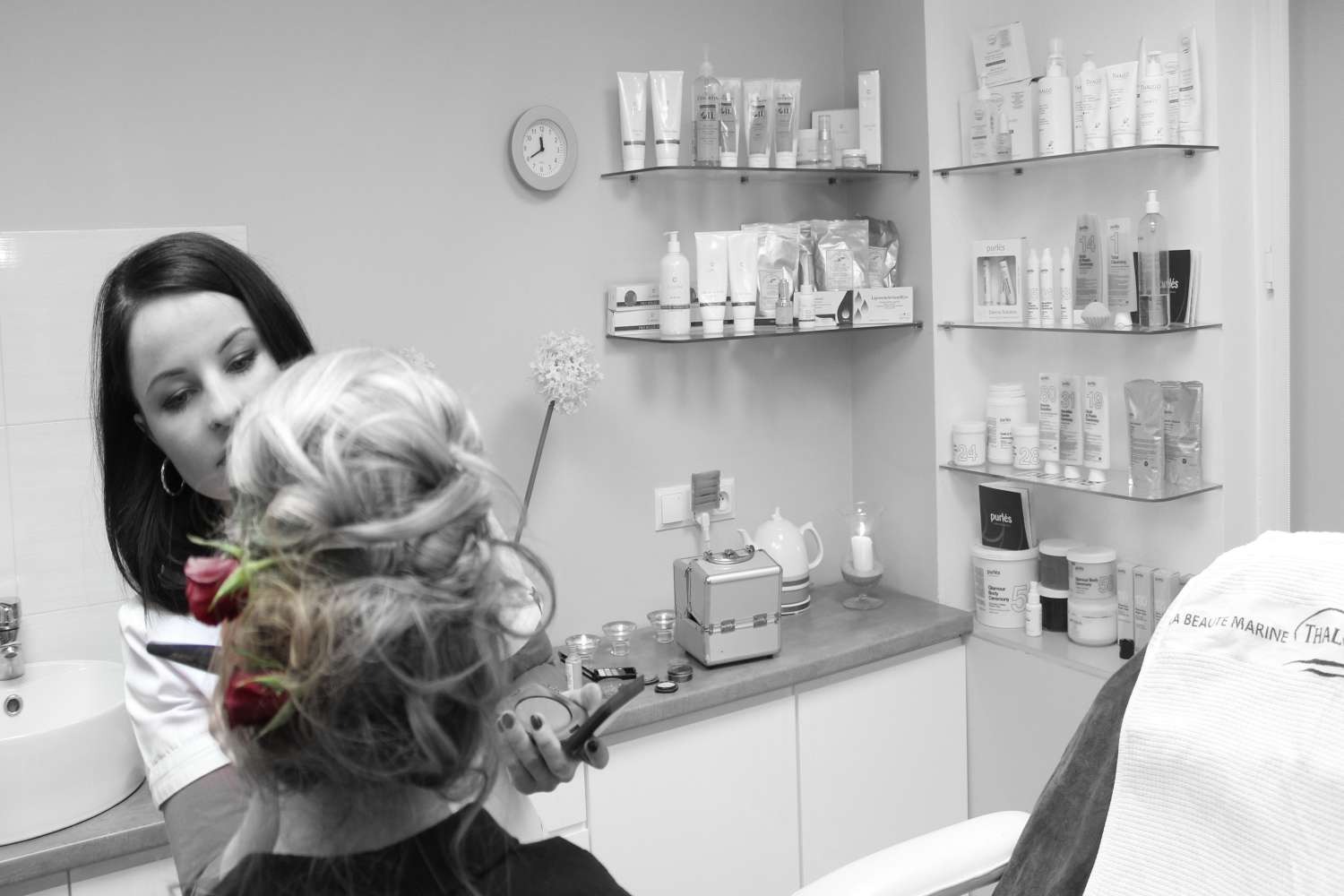 Fryzjerka upina włosy klientki z czerwonymi różami, w tle półki z kosmetykami i akcesoriami w sterylnym, jasnym wnętrzu.