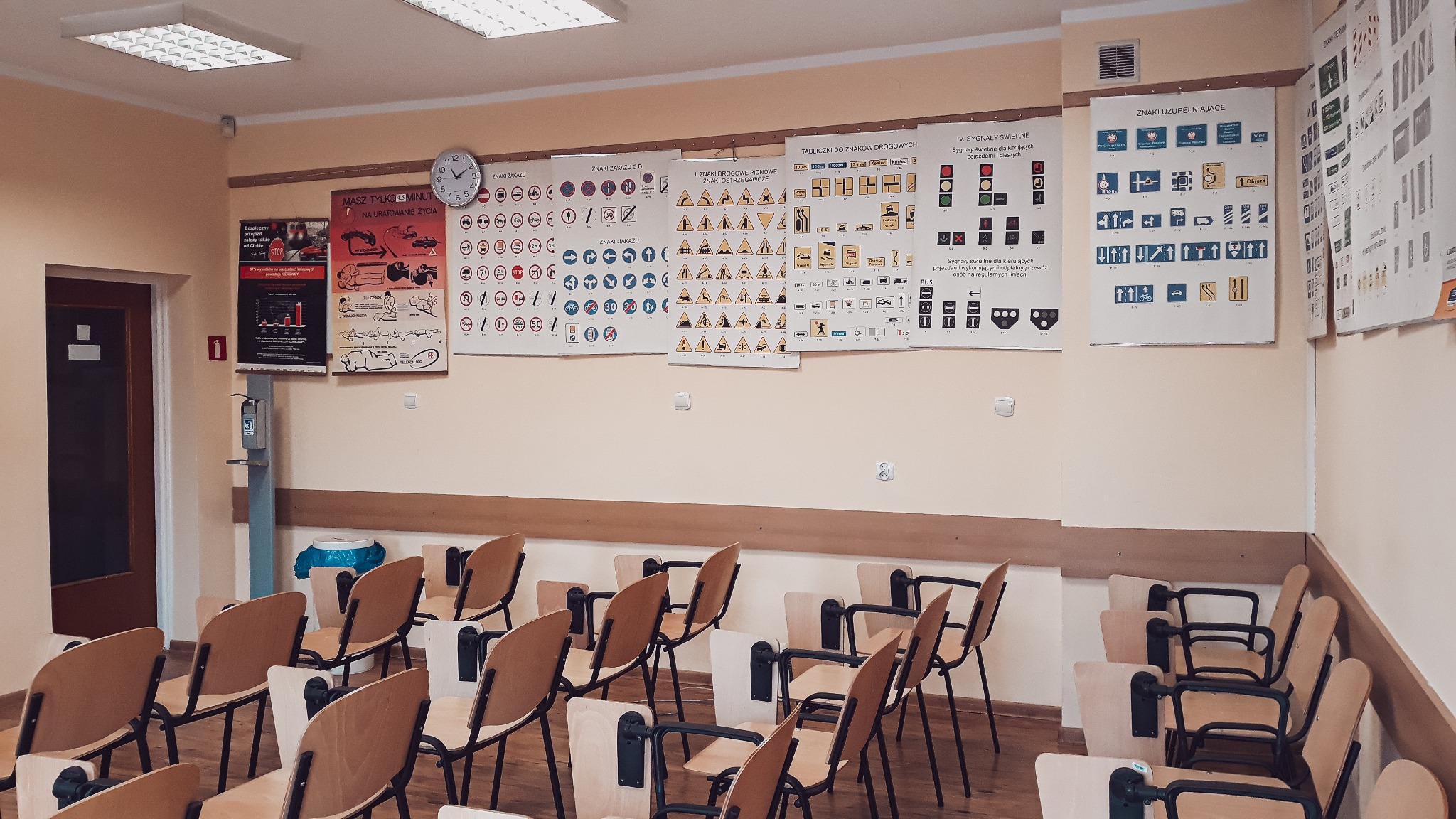 Sala wykładowa z krzesłami i plakatami ze znakami drogowymi na ścianach, zegar na ścianie, dozownik do dezynfekcji przy wejściu.