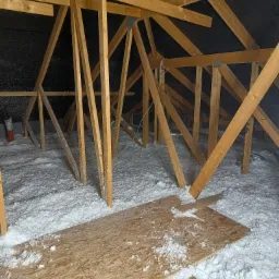 Poddasze z widoczną drewnianą konstrukcją dachu i warstwą białej izolacji, fragment płyty OSB na podłodze, czarna folia dachowa w tle.