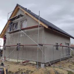 Prace elewacyjne w Oławie: dom jednorodzinny w trakcie nakładania tynku zewnętrznego, widoczne rusztowania, pracownicy oraz materiały budowlane na ziemi.