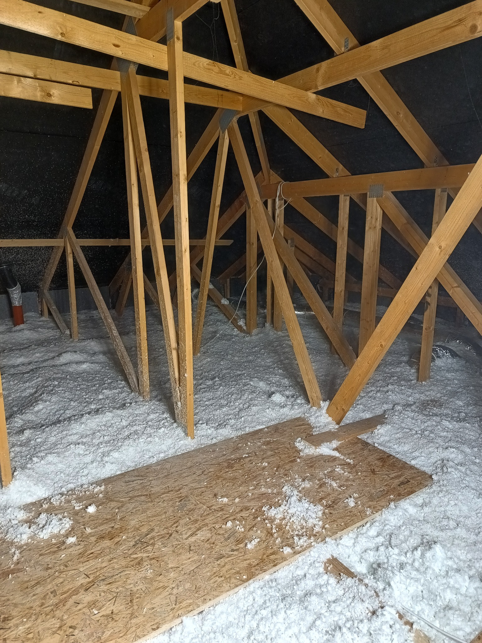 Poddasze z widoczną drewnianą konstrukcją dachu i warstwą białej izolacji, fragment płyty OSB na podłodze, czarna folia dachowa w tle.