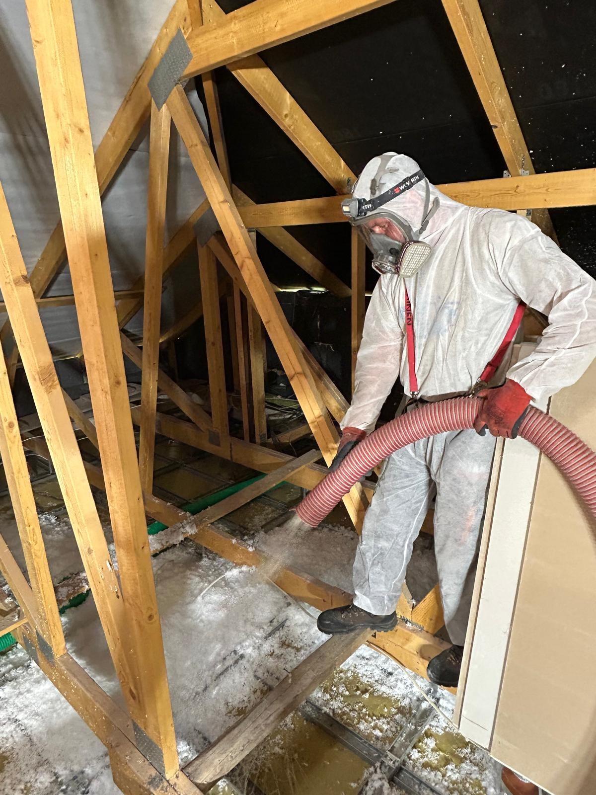 Pracownik w kombinezonie ochronnym i masce, aplikuje izolację z włókien na poddaszu, używając czerwonej rury i specjalistycznego sprzętu, widoczne drewniane elementy konstrukcji dachu.