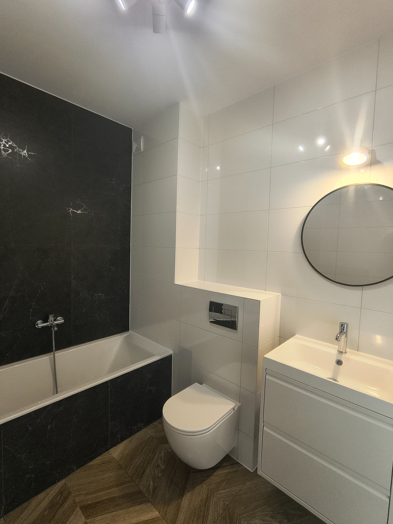 Nowoczesna łazienka z czarno-białymi płytkami, drewnianą podłogą w jodełkę i okrągłym lustrem z podświetleniem. Minimalistyczny design, armatura chromowana.