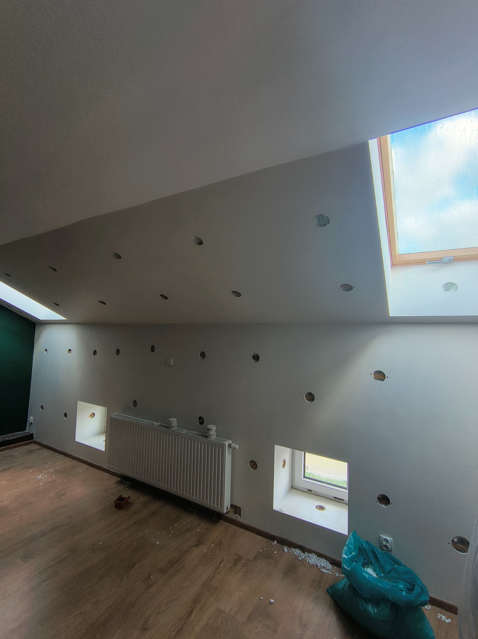 Poddasze po montażu płyt gipsowych, widoczne otwory na oświetlenie, okna dachowe i grzejnik. Na podłodze resztki materiałów budowlanych i worki na śmieci.