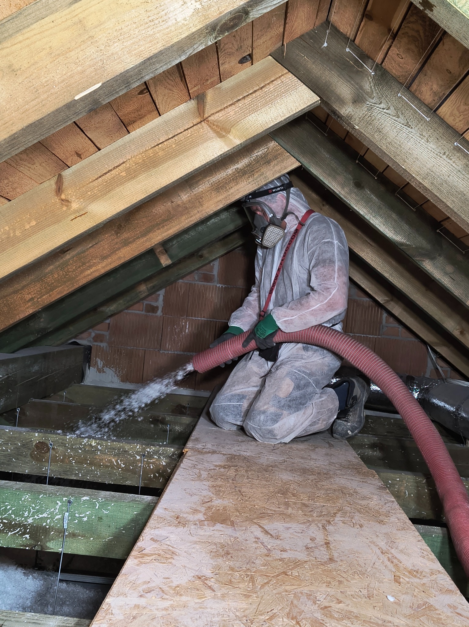 Pracownik w kombinezonie i masce natryskuje izolację na poddaszu, widoczne drewniane elementy konstrukcji dachu i ceglana ściana. Czerwony wąż i biała piana izolacyjna.