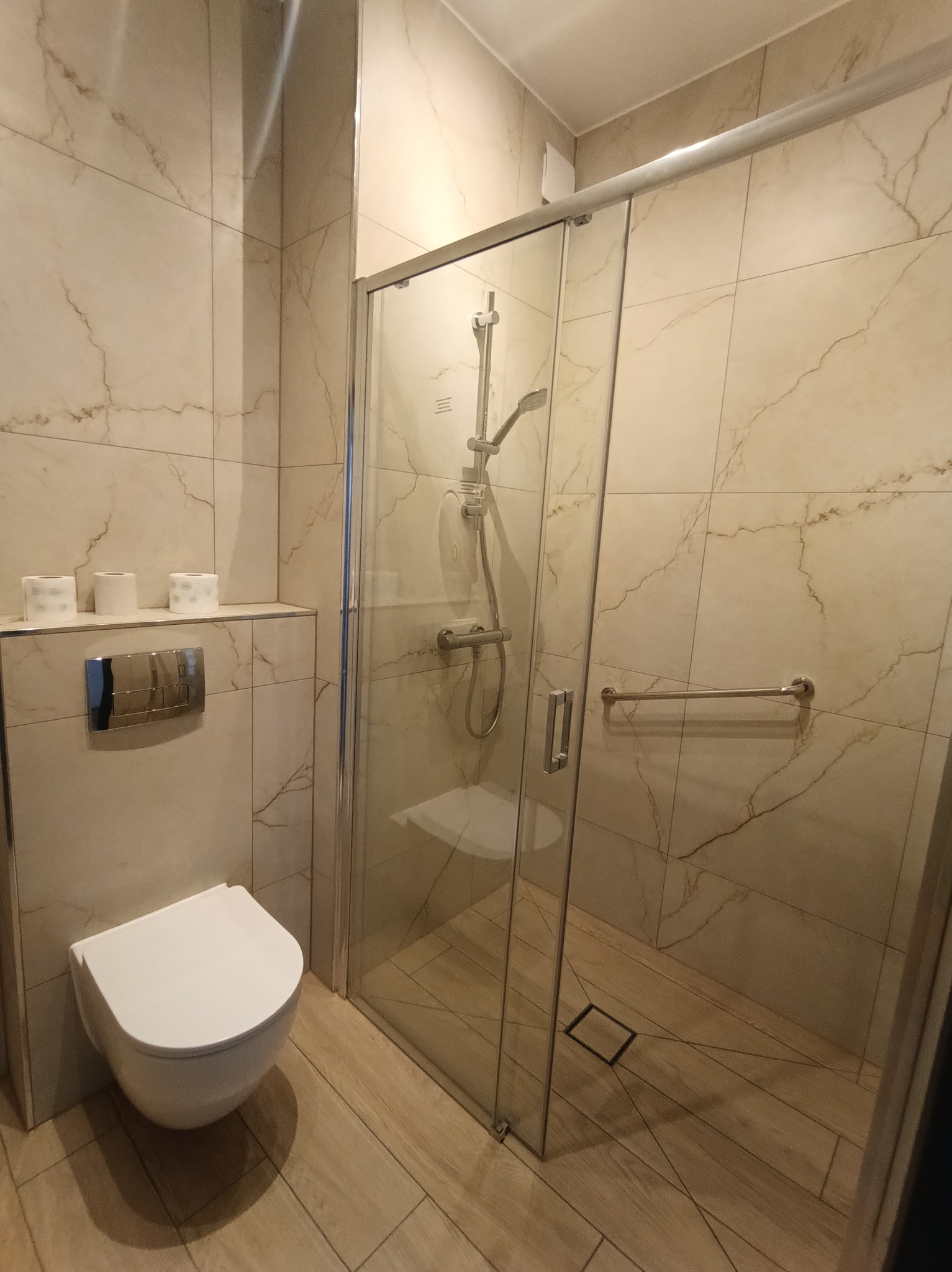 Nowoczesna łazienka z kabiną prysznicową typu walk-in, wykończona płytkami imitującymi marmur. Widoczny podwieszany sedes i minimalistyczna armatura.