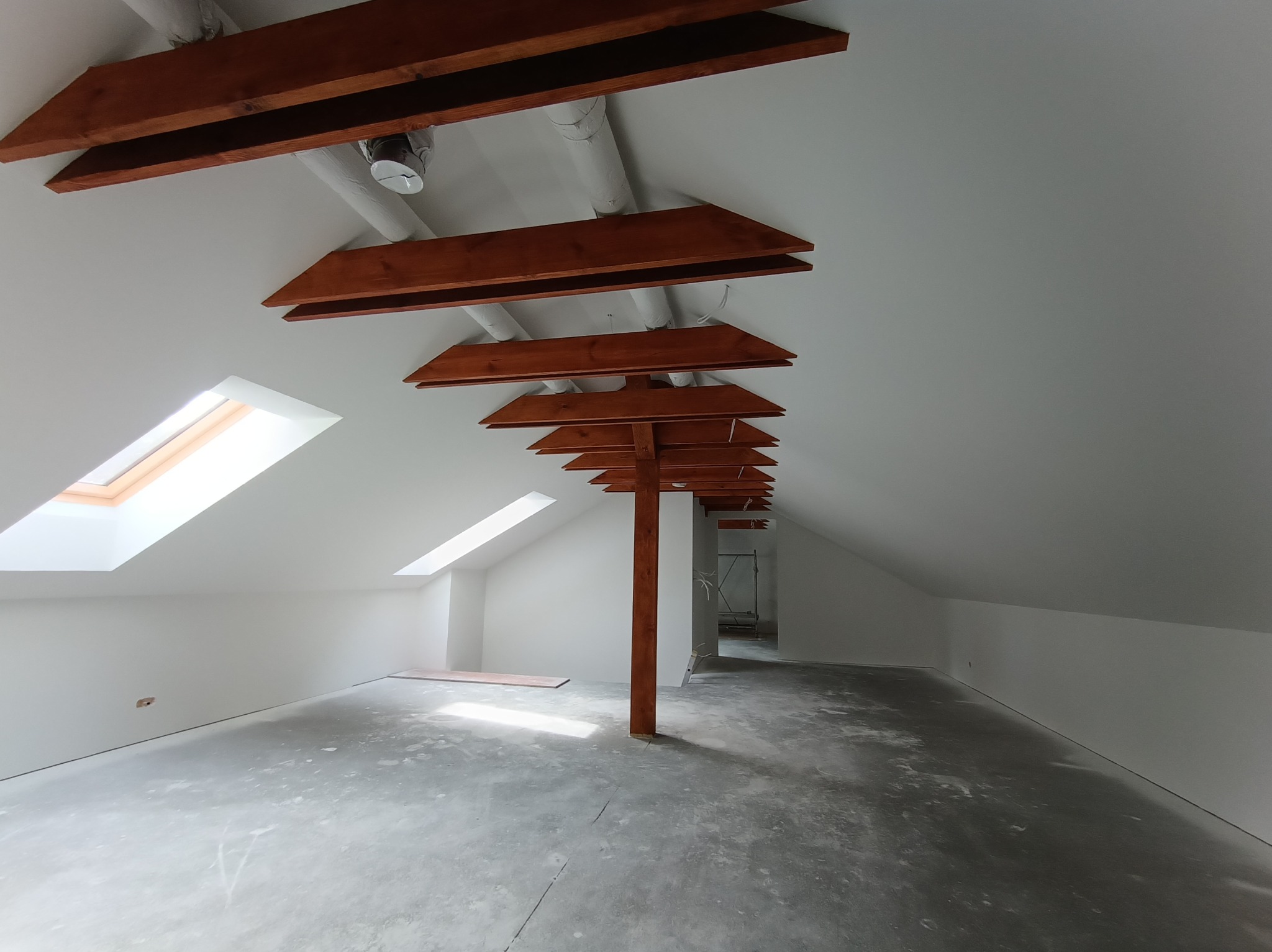 Poddasze po remoncie: białe ściany, okna dachowe, drewniane elementy konstrukcyjne. Surowa podłoga, widoczne instalacje. Jasne, przestronne wnętrze w trakcie wykańczania.