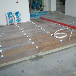 Układanie płytek imitujących drewno na podłodze, z użyciem poziomicy, krzyżyków dystansowych i klipsów poziomujących, worki z zaprawą w tle.