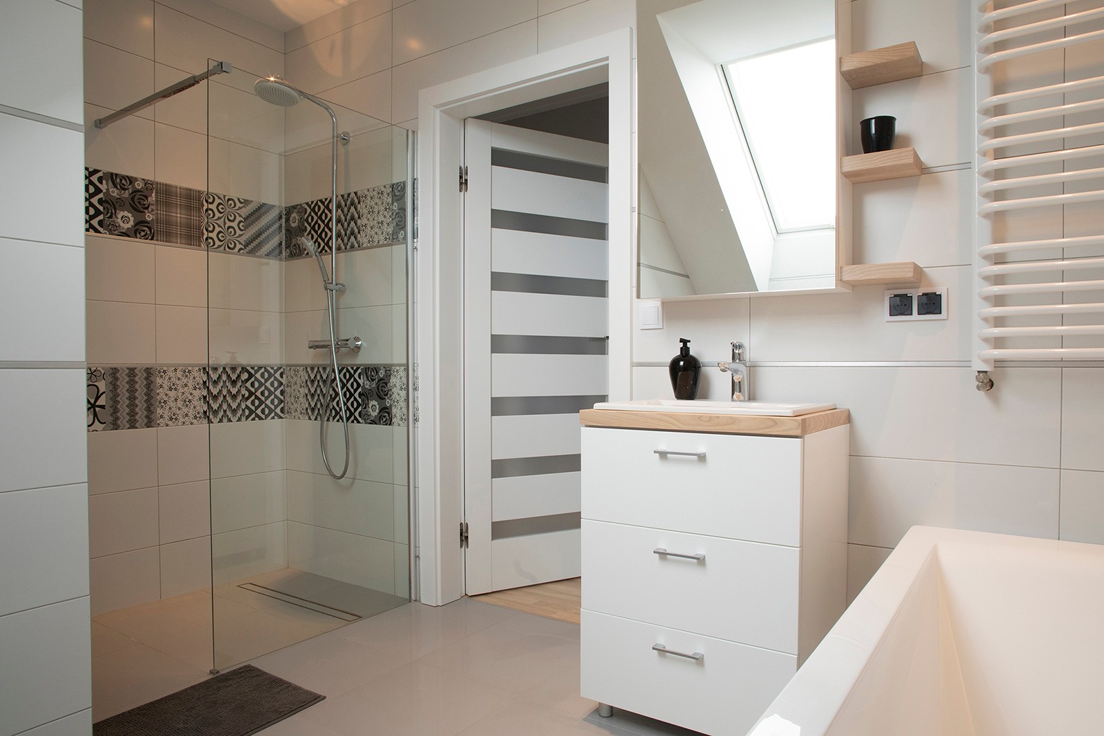 Jasna łazienka z kabiną prysznicową z dekoracyjnymi płytkami, białą szafką z drewnianym blatem, lustrem z półkami i grzejnikiem drabinkowym.