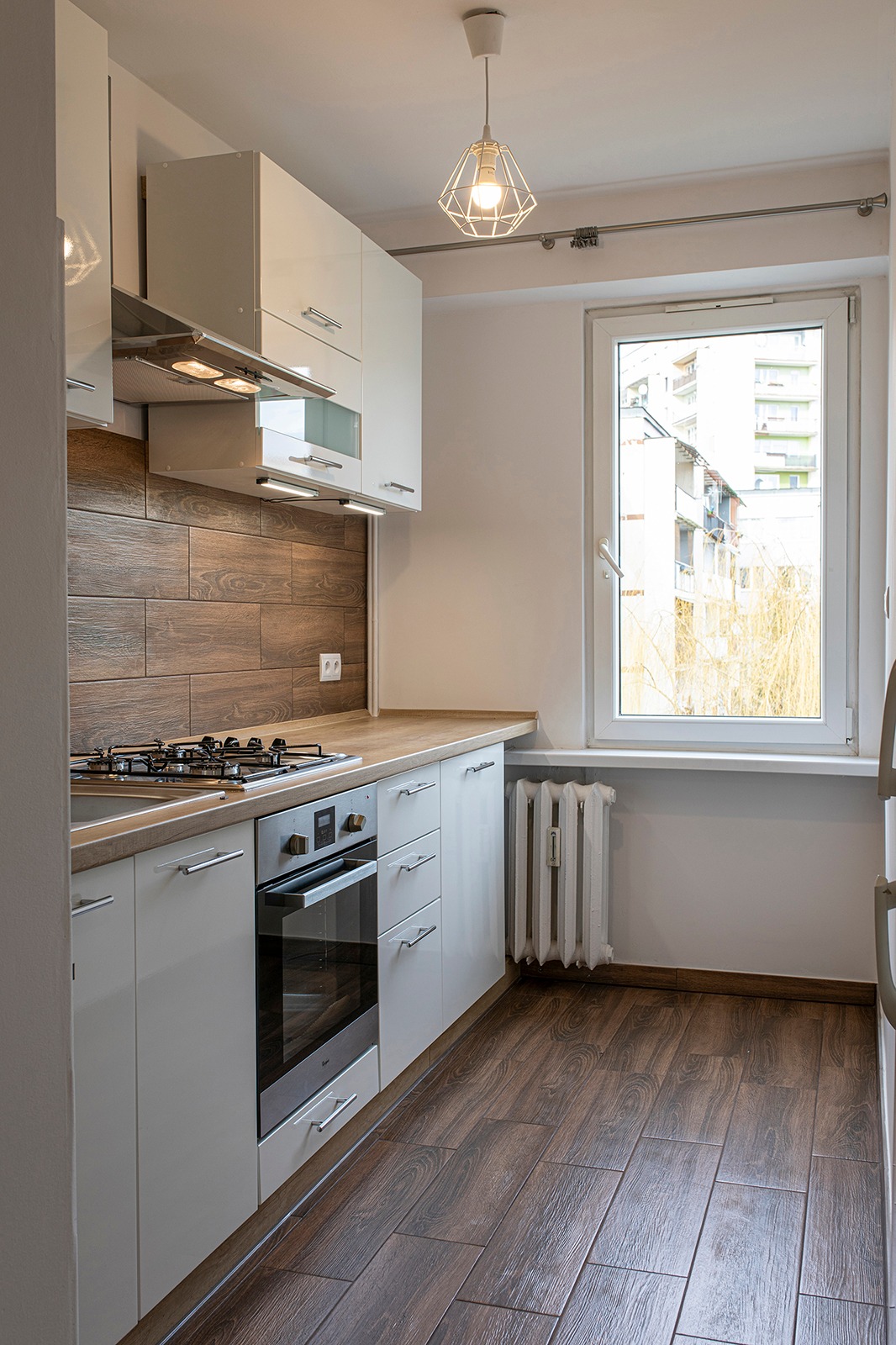 Nowoczesna, jasna kuchnia z białymi, lakierowanymi szafkami, drewnopodobnymi panelami ściennymi i podłogą, widziana z perspektywy wejścia, z oknem z widokiem na drzewa bez liści.