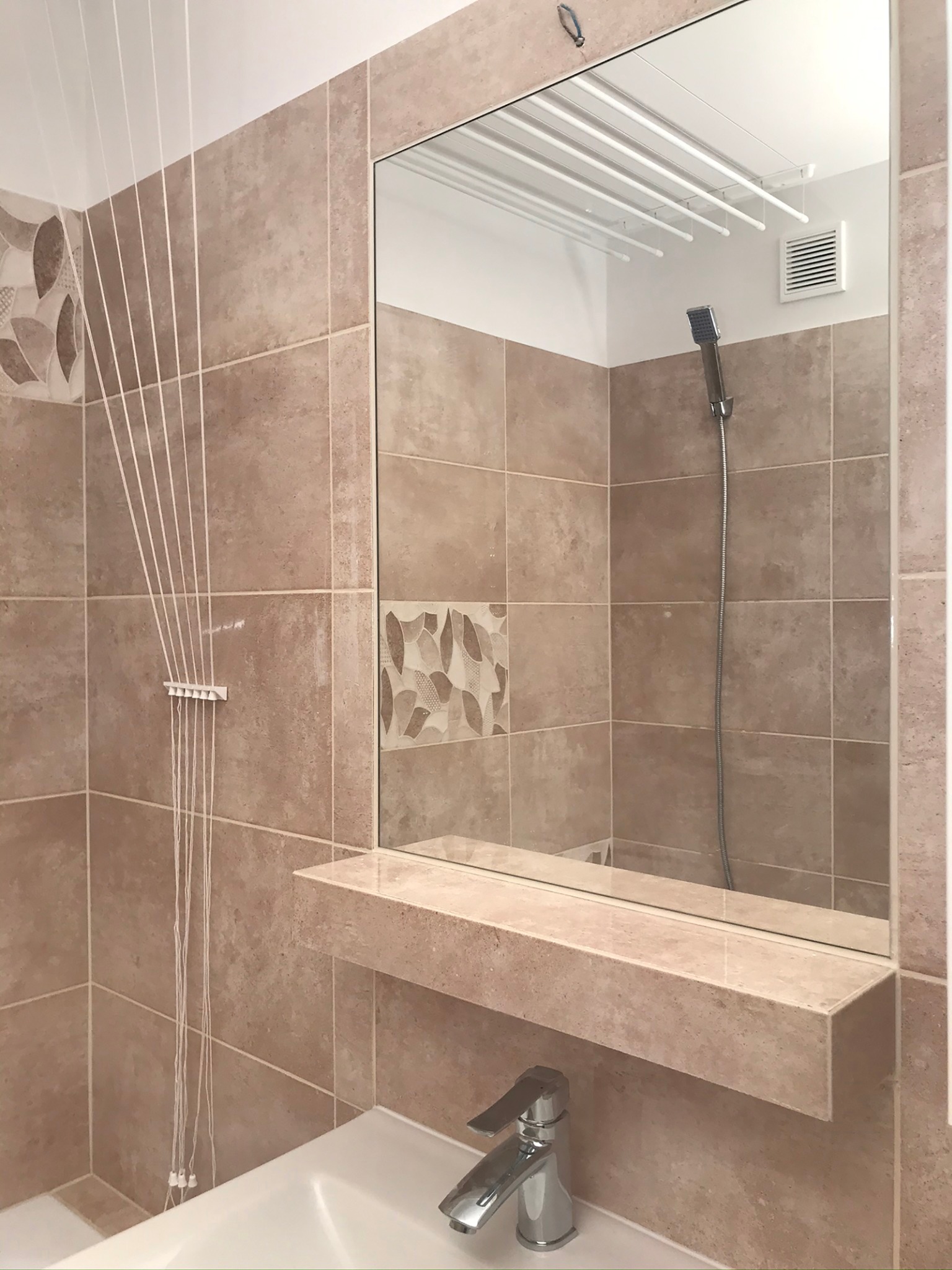 Ukończona łazienka z beżowymi płytkami, wisząca suszarka na pranie, lustro z półką nad umywalką i widocznym odbiciem kabiny prysznicowej.