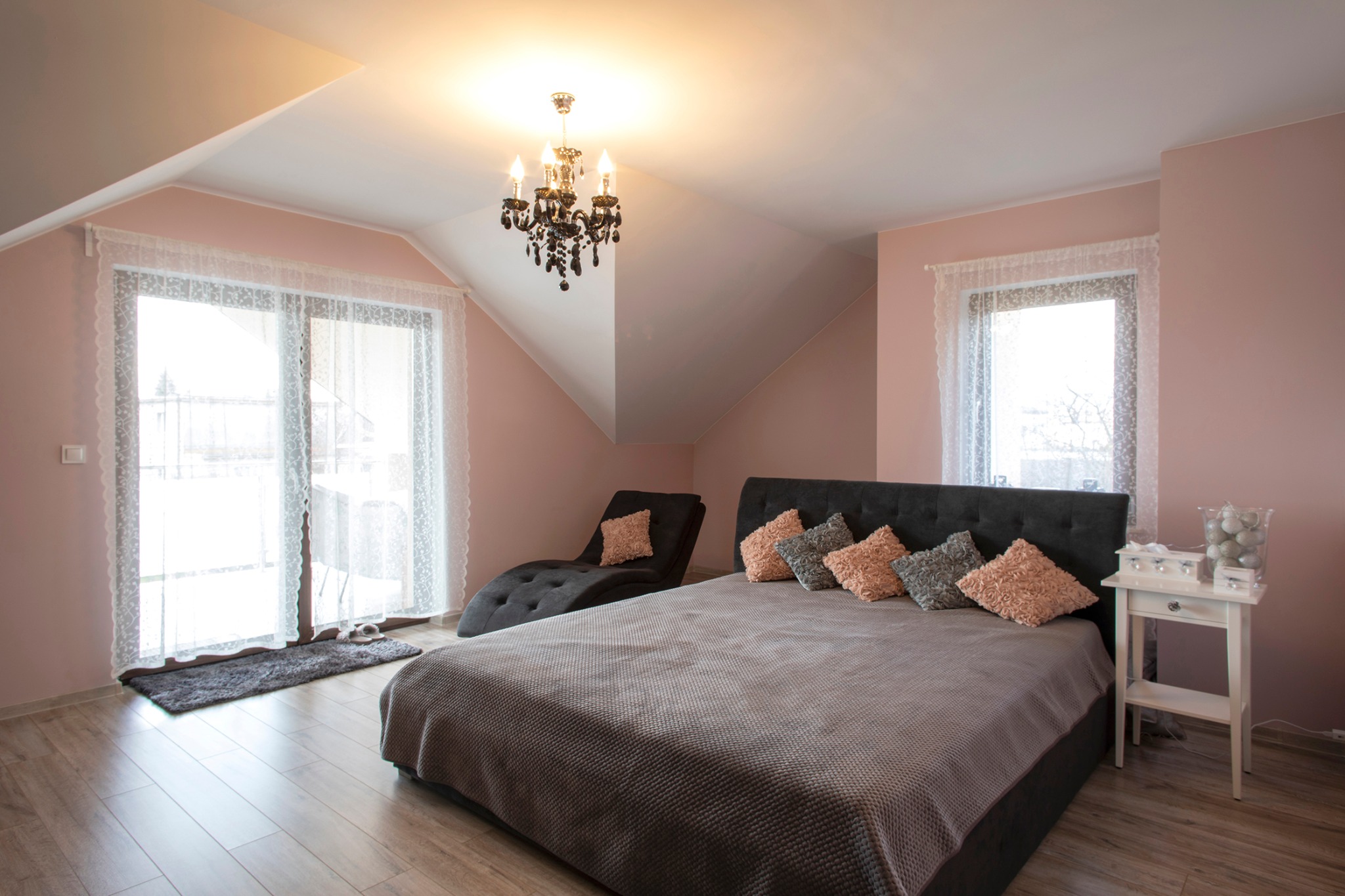 Jasny pokój na poddaszu z łóżkiem, szezlongiem i żyrandolem, ściany pomalowane na pudrowy róż.