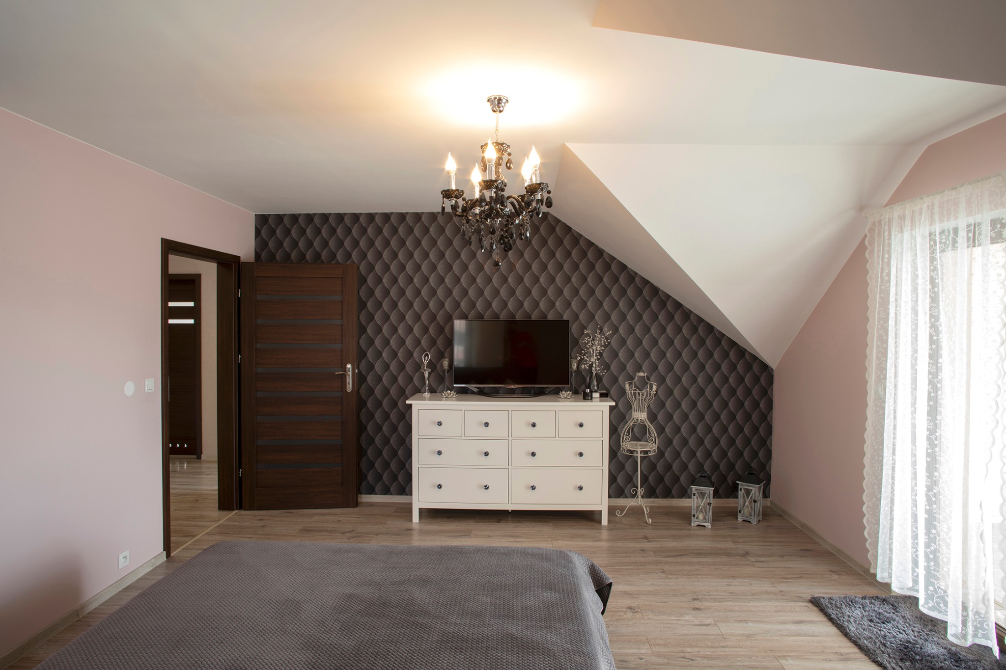 Jasny pokój z łóżkiem, komodą pod telewizorem i ciemną tapetą w romby, oświetlony ozdobnym żyrandolem.
