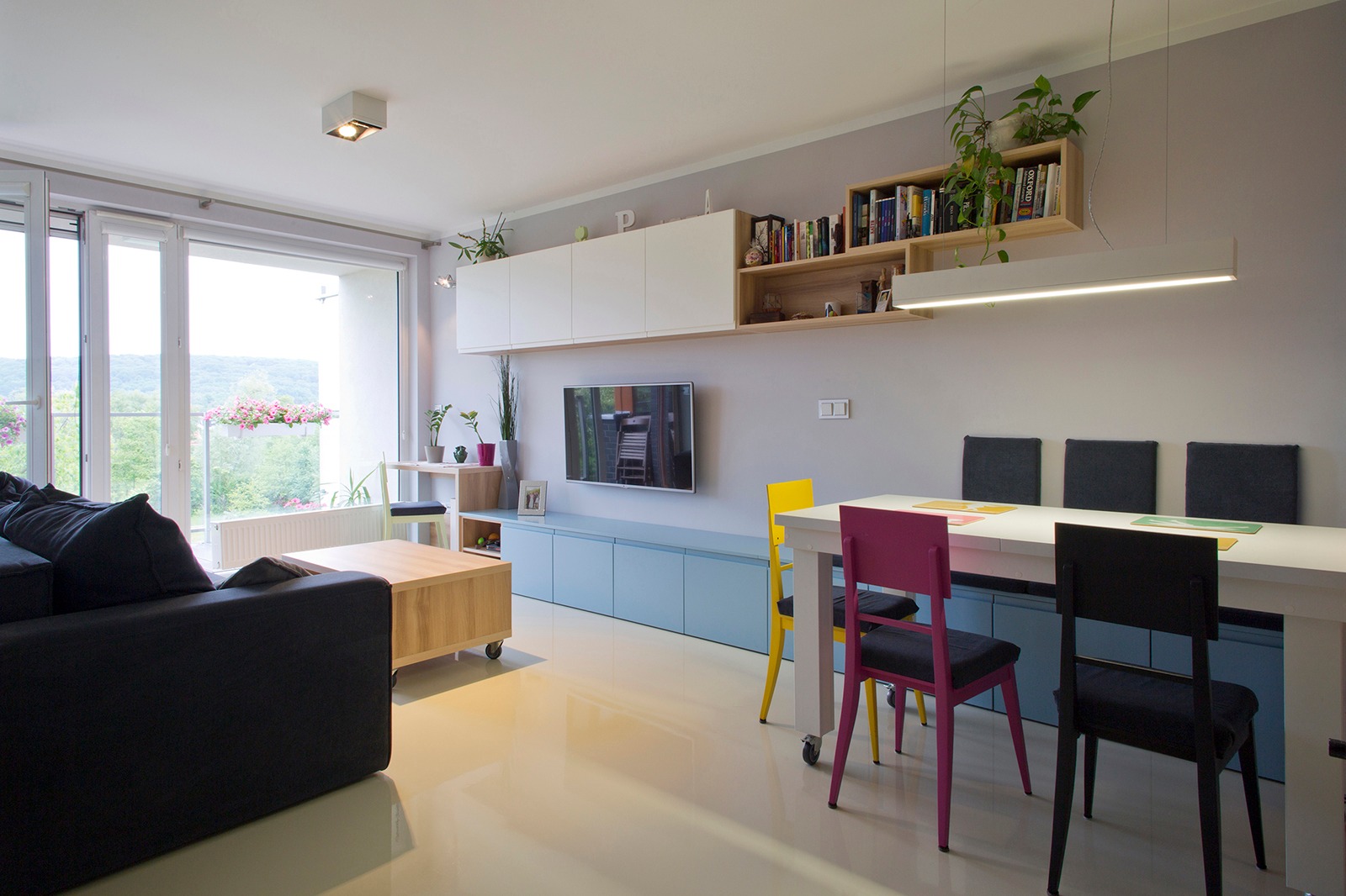 Nowoczesny salon z białymi szafkami wiszącymi, drewnianymi półkami z książkami i roślinami, niebieską szafką RTV, telewizorem, stołem z kolorowymi krzesłami i dużym oknem balkonowym z widokiem...