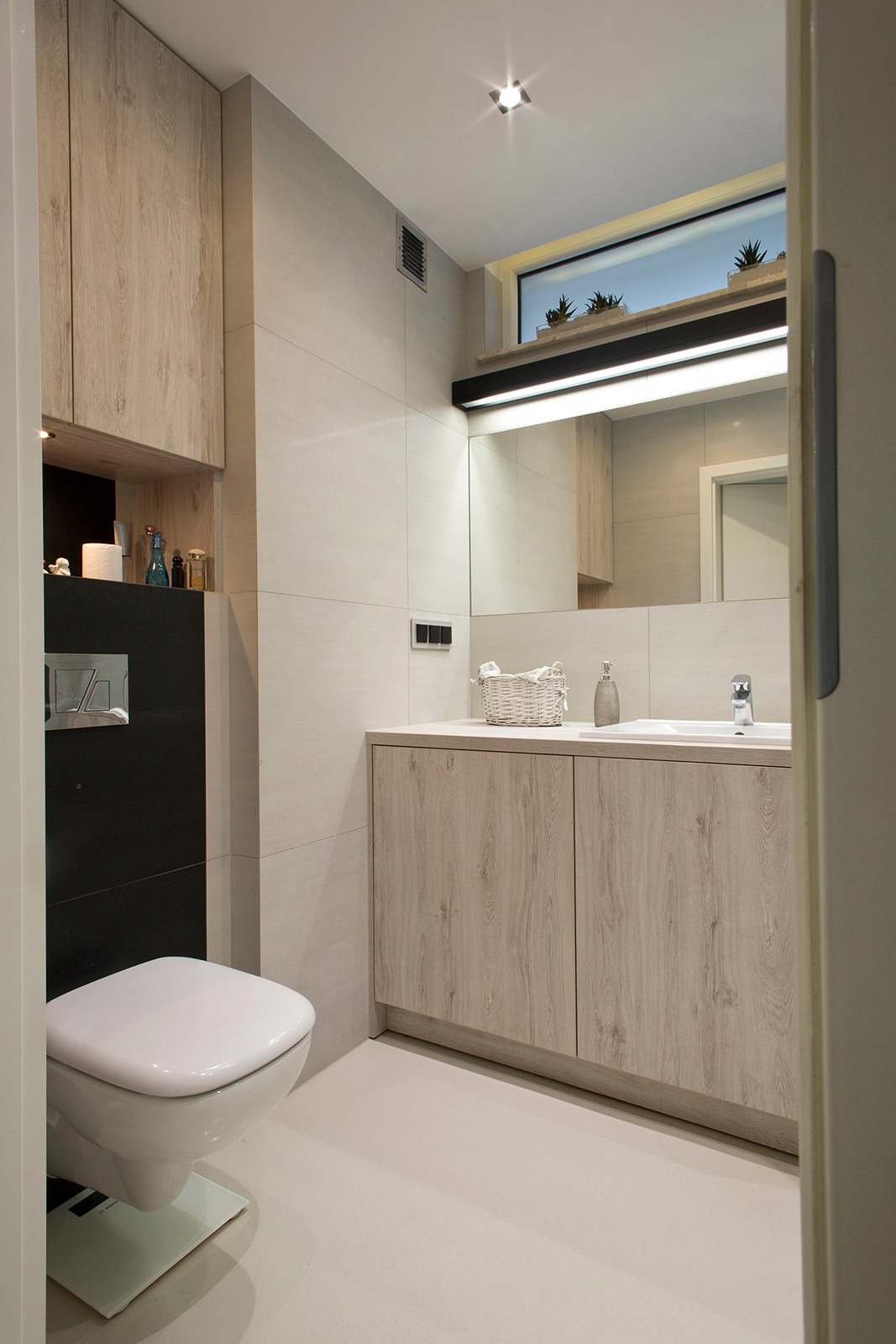 Nowoczesna łazienka z drewnopodobnymi szafkami, wiszącą toaletą na szklanej podstawce, dużym lustrem i oknem z roślinami doniczkowymi, oświetlona punktowym światłem.