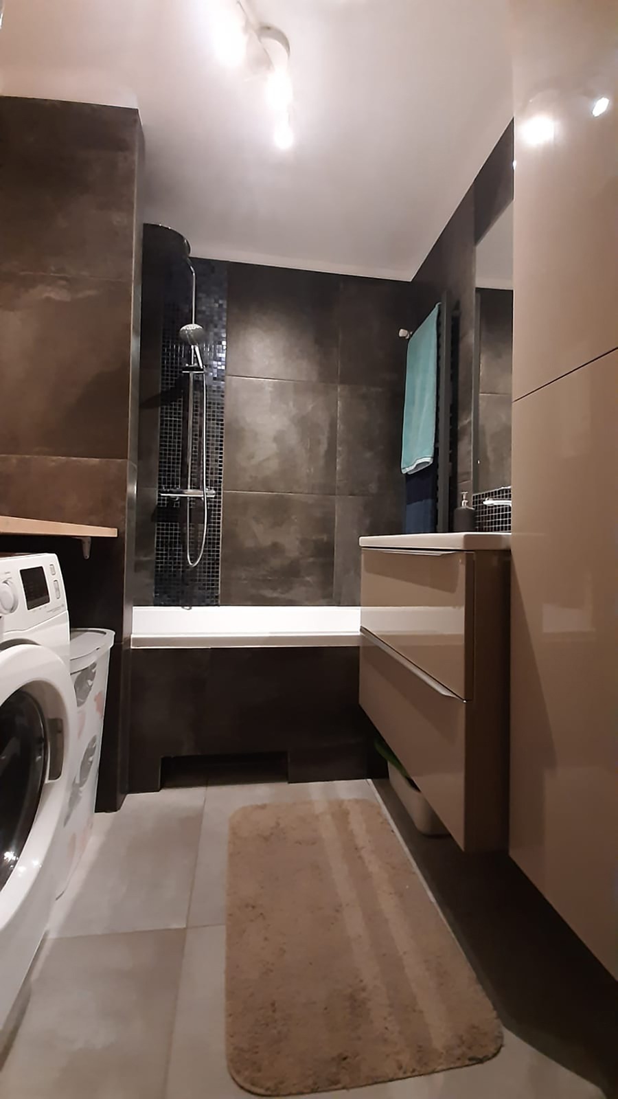 Nowoczesna łazienka z brązowymi płytkami, kabiną prysznicową typu walk-in z mozaiką, beżową szafką umywalkową, pralką i beżowym dywanikiem.