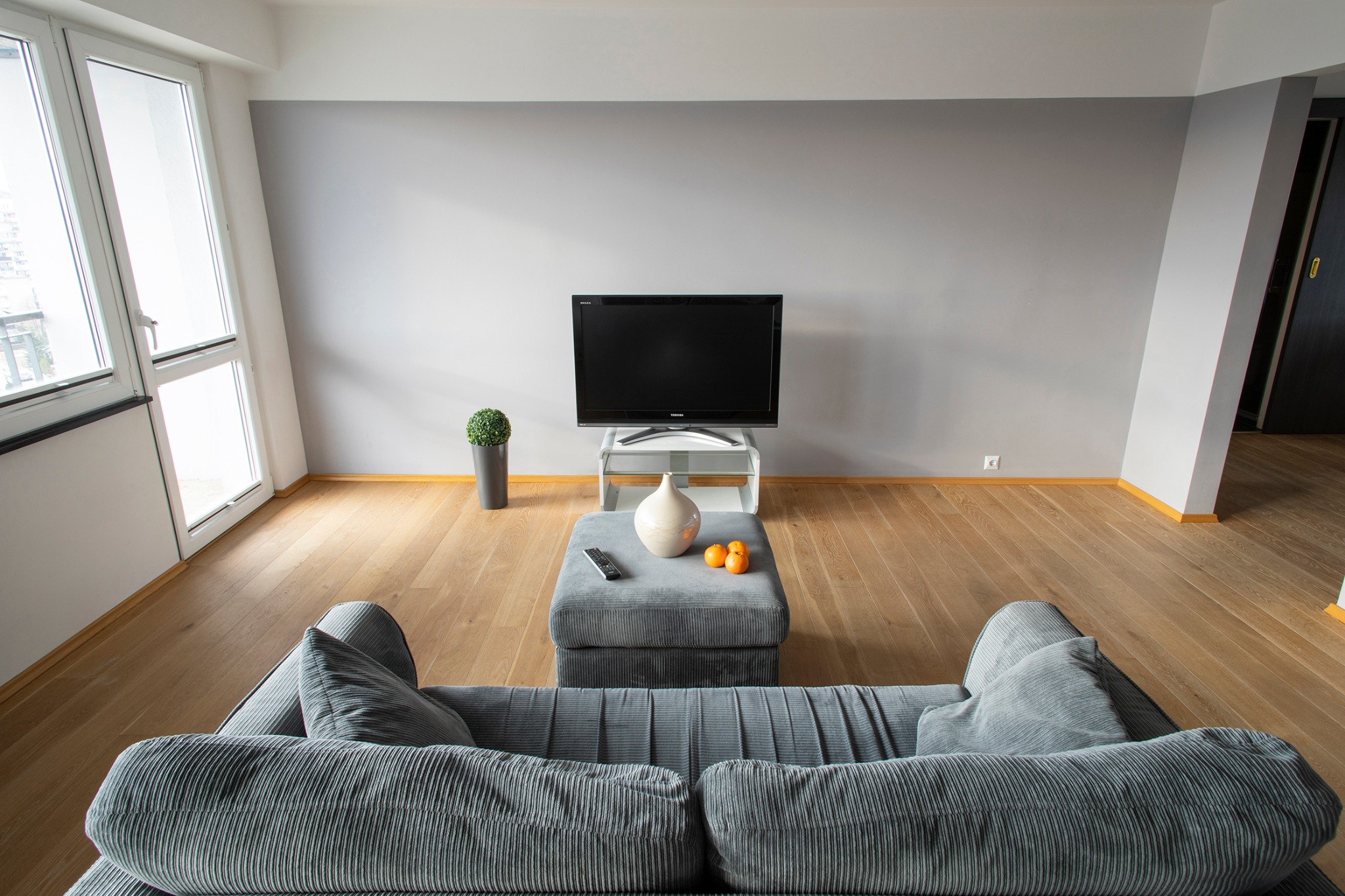 Nowoczesny salon z szarą ścianą, drewnianą podłogą, telewizorem, szarą sofą i pufą, z dekoracjami w postaci wazonu, mandarynek i rośliny doniczkowej.