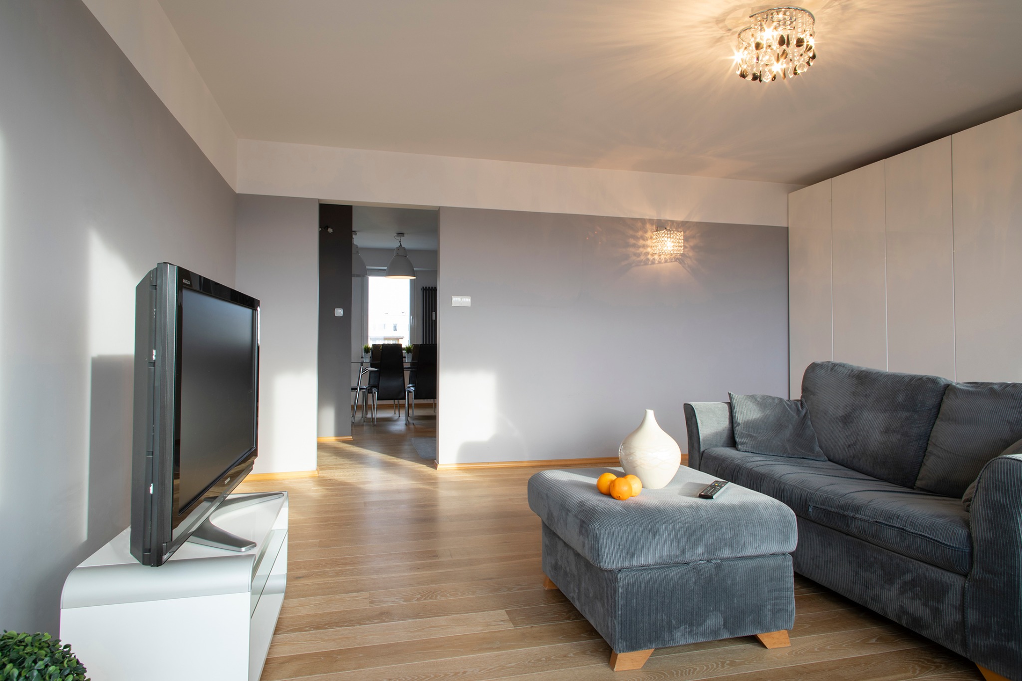 Jasne wnętrze salonu z szarą sofą z podnóżkiem, telewizorem na białej szafce RTV i drewnianą podłogą; w tle widoczna jadalnia