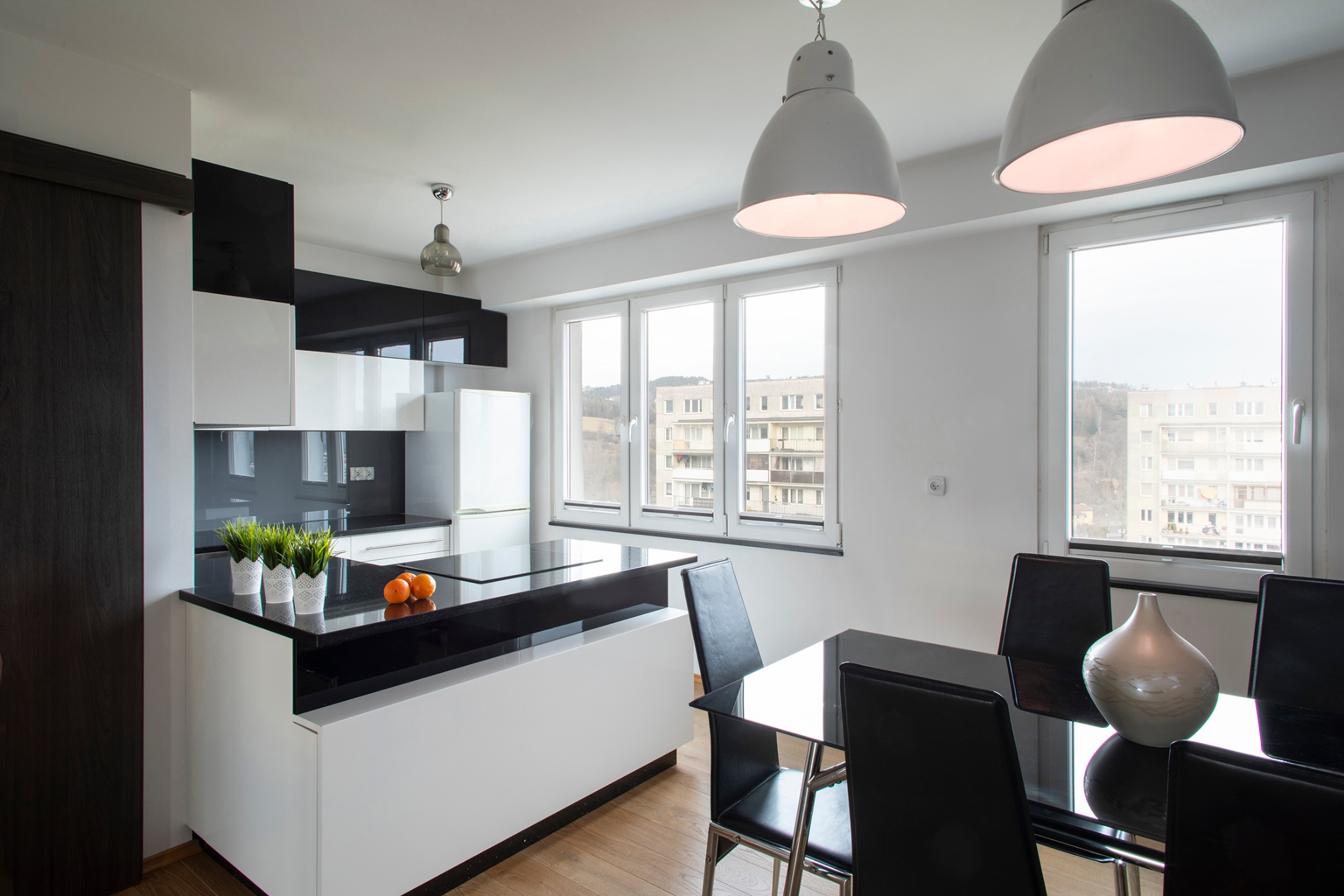 Nowoczesna kuchnia z czarno-białymi szafkami, czarnym blatem, drewnianą podłogą i stołem z krzesłami, oświetlona lampami w stylu industrialnym, z widokiem na budynki za oknem.
