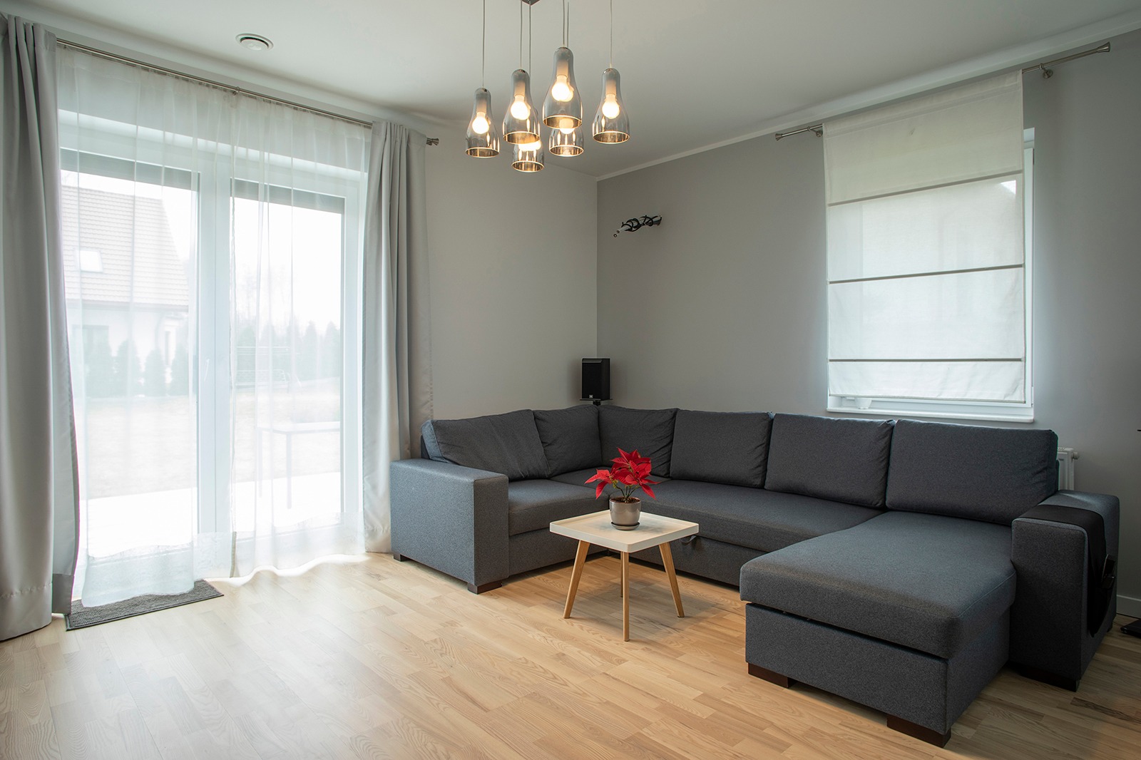 Jasne wnętrze salonu po remoncie: szary narożnik z czerwoną gwiazdą betlejemską na białym stoliku kawowym, parkiet, okno z zasłonami i roletą rzymską, nowoczesna lampa wisząca.