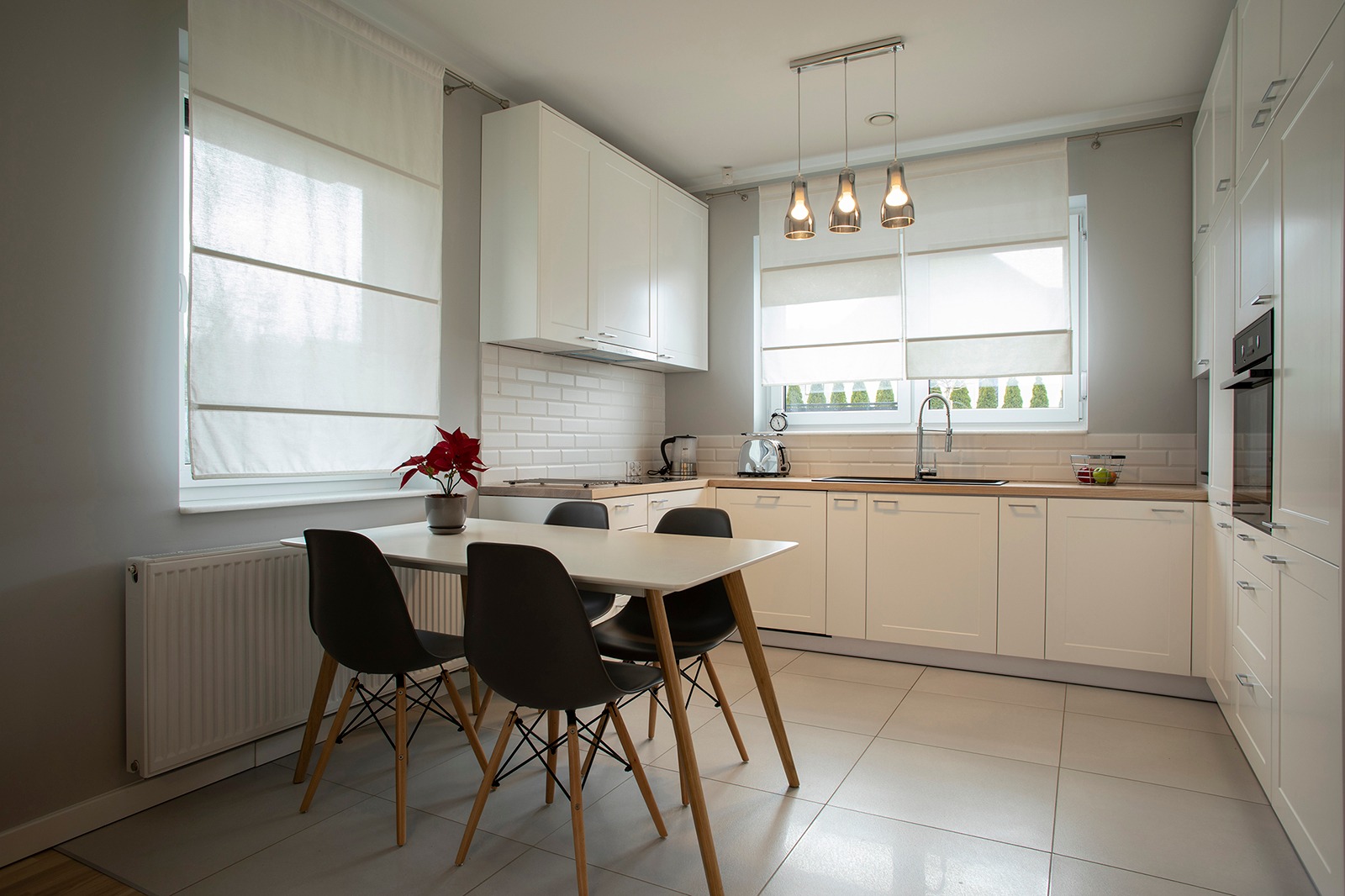 Jasna kuchnia z białymi szafkami, drewnianym blatem, szarą ścianą i podłogą w płytkach, z białym stołem i czarnymi krzesłami. Na stole stoi czerwona gwiazda betlejemska w szarej doniczce.