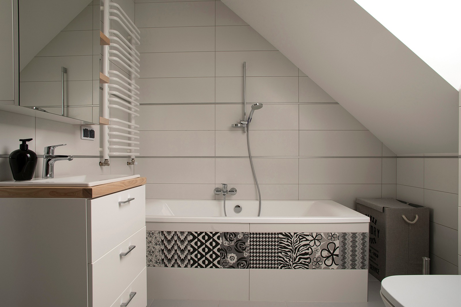 Biała łazienka na poddaszu ze skośnym sufitem, z wanną obłożoną dekoracyjnymi płytkami w geometryczne i kwiatowe wzory, białą szafką z drewnianym blatem i czarnym dozownikiem na mydło.