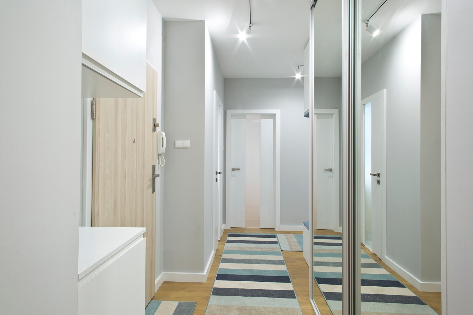 Jasne, nowoczesne wnętrze przedpokoju z drewnianą podłogą, szarą ścianą, białymi drzwiami wewnętrznymi, lustrem w zabudowie i dywanem w niebieskie pasy.