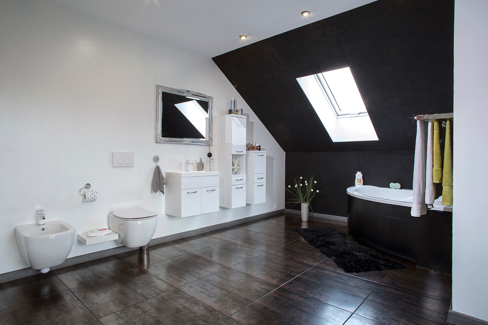 Nowoczesna łazienka na poddaszu z czarną ścianą, oknem dachowym, białymi meblami łazienkowymi i ciemną podłogą z płytek imitujących drewno.