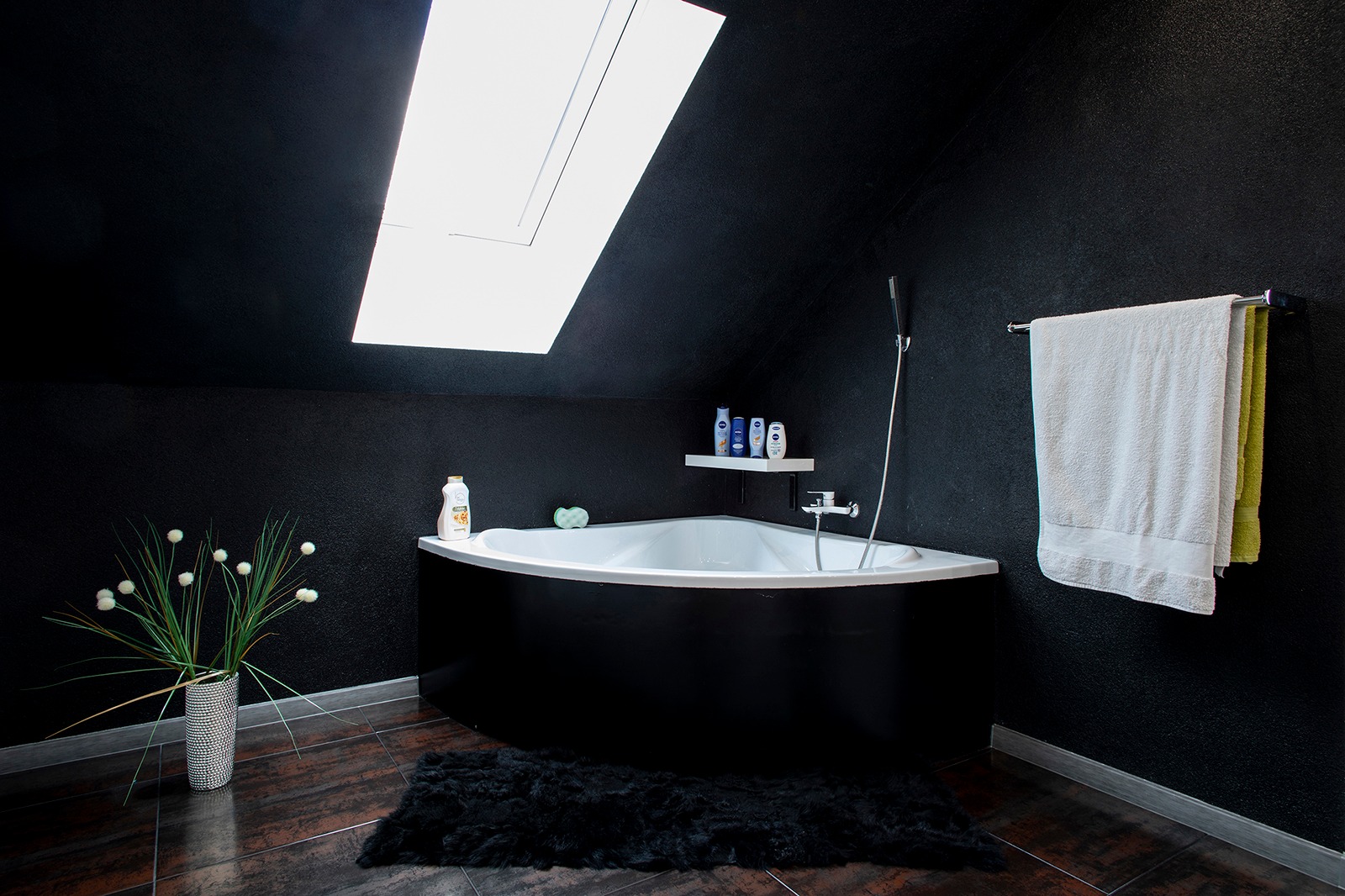 Czarno-biała łazienka na poddaszu z oknem dachowym, narożną wanną, ciemną podłogą, dekoracyjnym wazonem z kwiatami i puszystym czarnym dywanikiem.