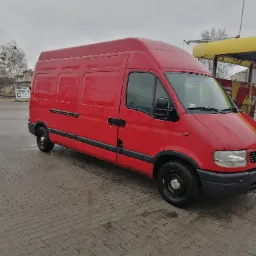 Czerwony bus dostawczy Opel Master zaparkowany na brukowanym placu, widoczny z boku pod kątem, z myjnią samochodową w tle.