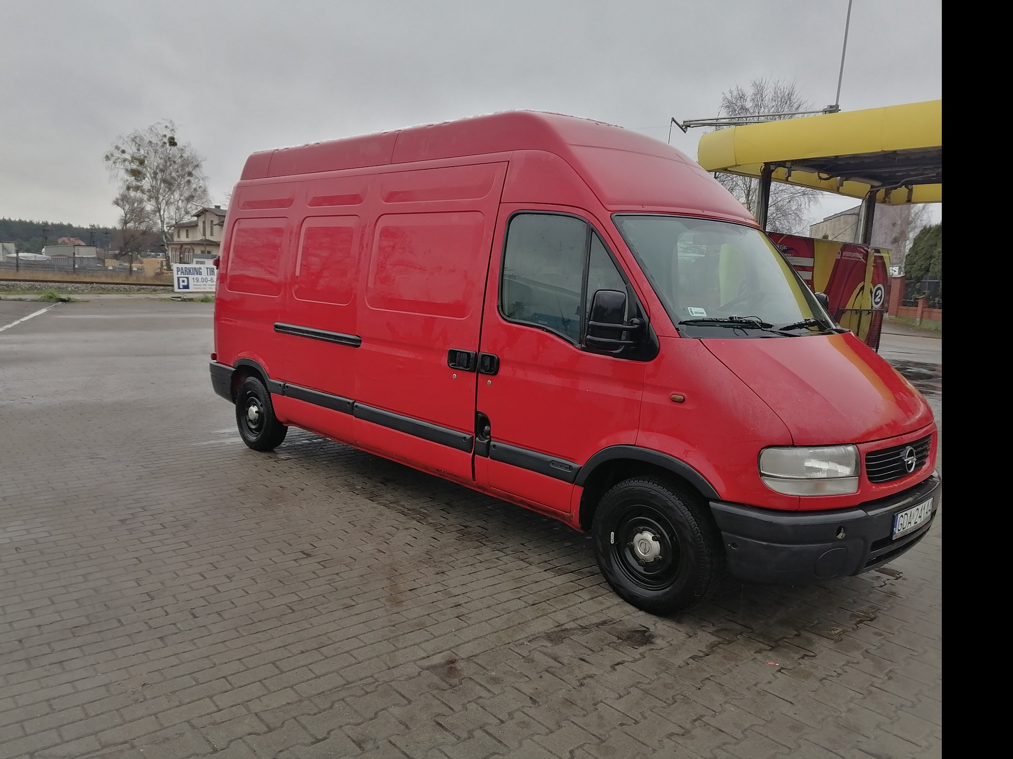 Czerwony bus dostawczy Opel Master zaparkowany na brukowanym placu, widoczny z boku pod kątem, z myjnią samochodową w tle.