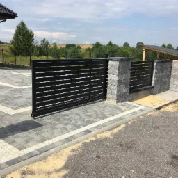 Nowoczesna, pozioma brama wjazdowa z czarnych profili, osadzona między dwoma szarymi, murowanymi słupkami z dekoracyjną fakturą, na tle kostki brukowej i zieleni.
