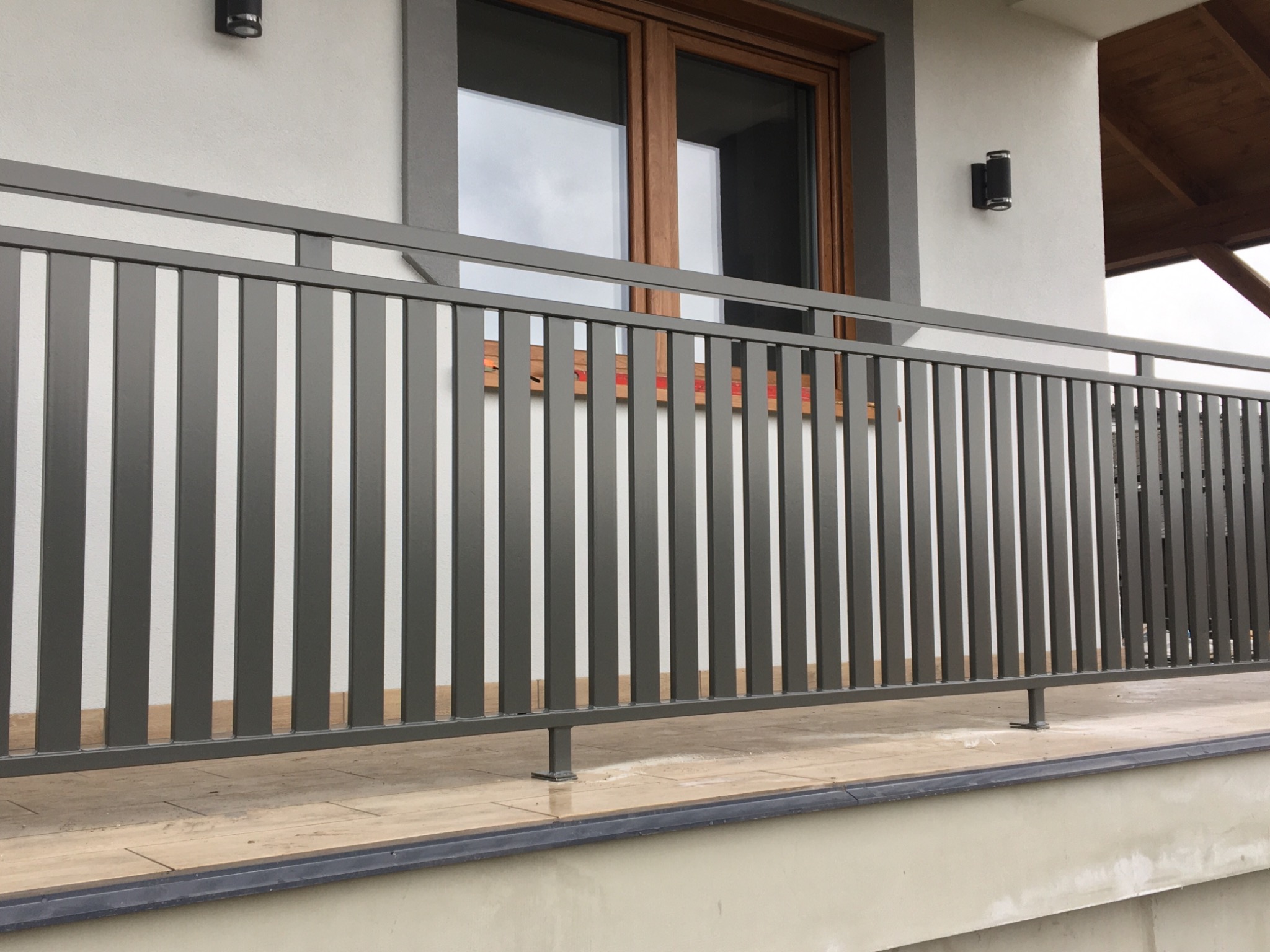 Szare metalowe balustrady balkonowe z pionowymi szczebelkami przed oknem z drewnianą ramą i zewnętrznymi lampami ściennymi.